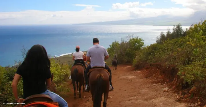 Riders on horseback following a dirt trail toward the ocean on Maui